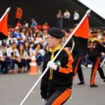 Desbravadores em desfile: Grupo de jovens Desbravadores marchando em formação, vestidos com uniformes pretos e laranja, carregando bandeiras nas cores preto e laranja. Ao fundo, uma plateia de pessoas assistindo ao desfile. O ambiente sugere um evento ao ar livre, possivelmente um desfile ou celebração organizada por um clube de Desbravadores.