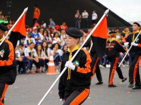 Desbravadores em desfile: Grupo de jovens Desbravadores marchando em formação, vestidos com uniformes pretos e laranja, carregando bandeiras nas cores preto e laranja. Ao fundo, uma plateia de pessoas assistindo ao desfile. O ambiente sugere um evento ao ar livre, possivelmente um desfile ou celebração organizada por um clube de Desbravadores.