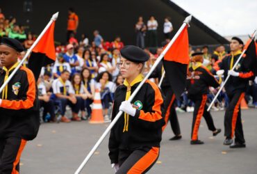 Desbravadores em desfile: Grupo de jovens Desbravadores marchando em formação, vestidos com uniformes pretos e laranja, carregando bandeiras nas cores preto e laranja. Ao fundo, uma plateia de pessoas assistindo ao desfile. O ambiente sugere um evento ao ar livre, possivelmente um desfile ou celebração organizada por um clube de Desbravadores.