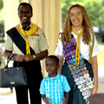 Três pessoas posando para uma foto em um ambiente ao ar livre com colunas ao fundo. À esquerda, um homem negro vestindo um uniforme escoteiro bege com faixa preta e gravata amarela, segurando uma bolsa preta. No centro, uma criança negra vestindo uma camisa xadrez azul clara. À direita, uma mulher de cabelos cacheados loiros vestindo um uniforme escoteiro branco com muitas insígnias coloridas e uma gravata amarela.